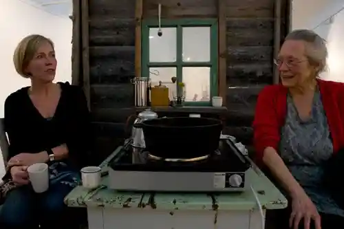 Two women sitting inside a rustic cabin setting, sharing a moment over coffee, with a stovetop and window visible in the background.