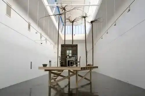 An installation featuring a raised wooden platform with barren trees and a small cabin, placed in a bright gallery space.