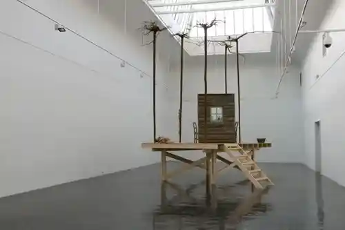 Wide-angle view of an art installation showcasing a wooden platform with a cabin and bare trees, illuminated by a skylight.